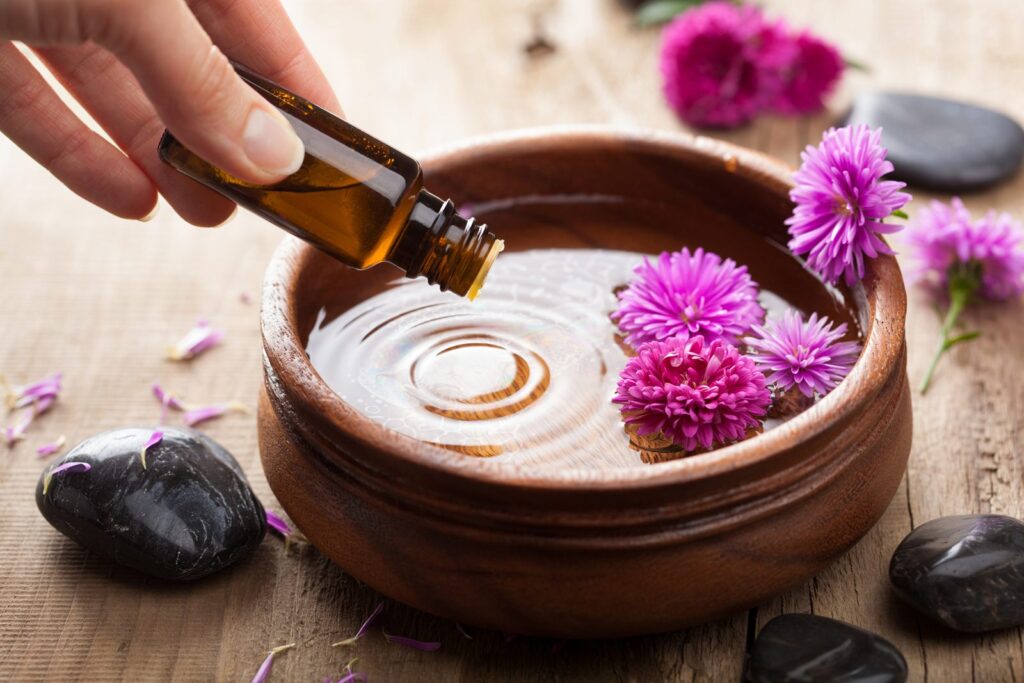 Essential oil being dropped from a dropper bottle into a wooden bowl of water surrounded by flowers