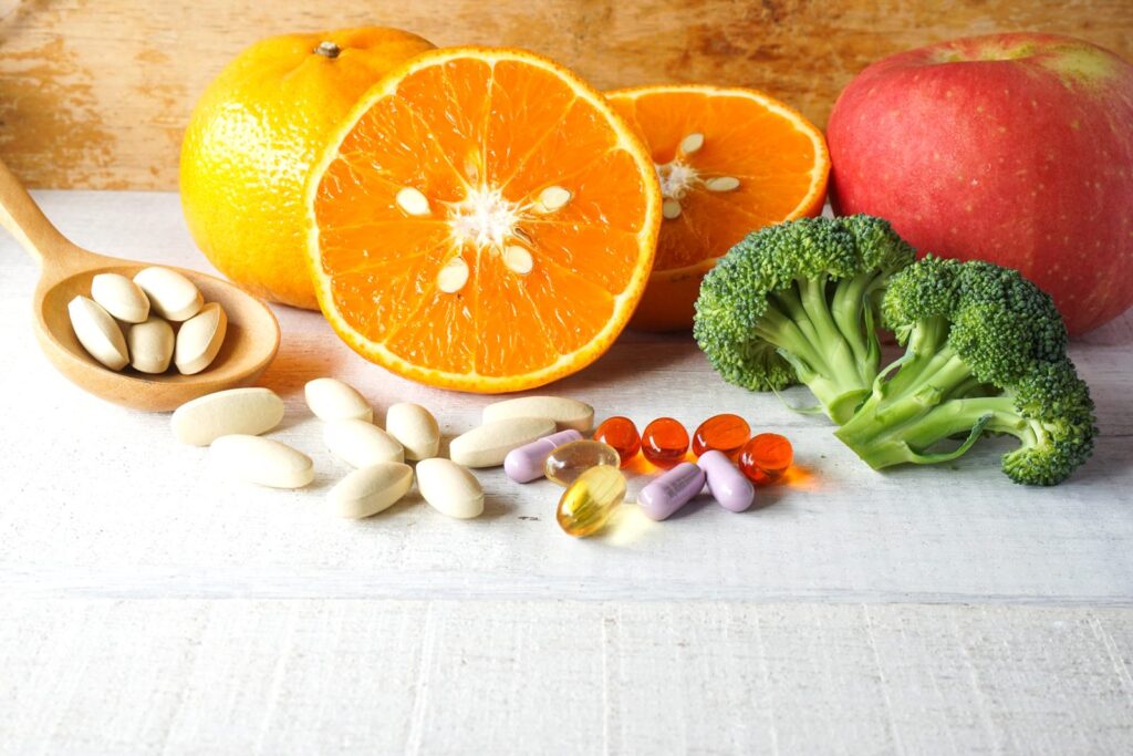 Vitamin tablets and capsules beside fresh oranges, an apple and broccoli